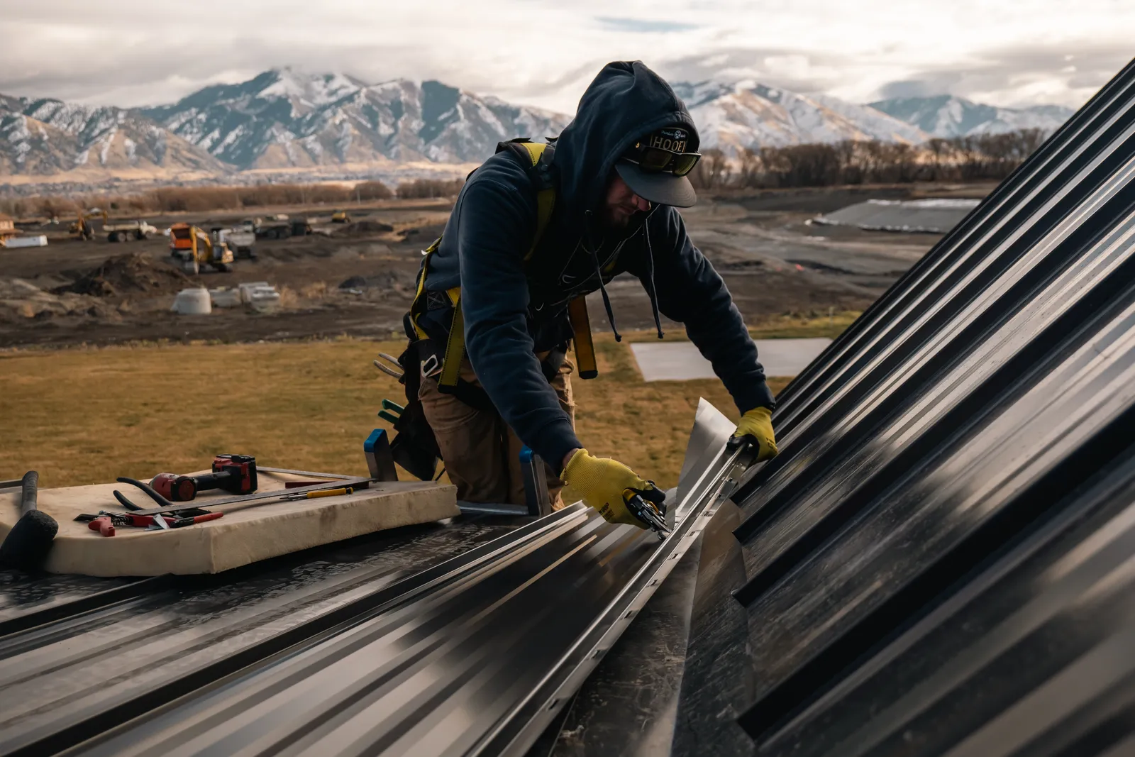 Homer Roofing installer hand-seaming standing seam metal panels with the Wasatch Mountains behind